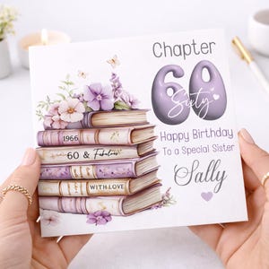 Può includere: Un biglietto d'auguri quadrato con una pila di cinque libri illustrati in tonalità viola e crema, decorato con fiori viola e farfalle. Il testo riporta Chapter 60, Sixty, Happy Birthday To a Special Sister, Sally, 1966, 60 & Fabulous e WITH LOVE.