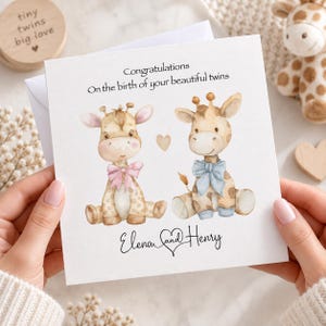 Op de afbeelding: Een vierkante wenskaart met een aquarelillustratie van twee babygiraffen die naast elkaar zitten. De linker giraffe draagt een roze strik en de rechter een blauwe strik. De tekst luidt: Congratulations On the birth of your beautiful twins Elena and Henry.