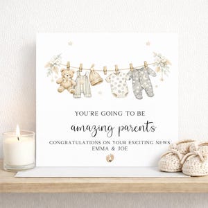 May include: A white greeting card with the text "YOU'RE GOING TO BE amazing parents". The card features illustrations of baby clothes hanging on a washing line, a teddy bear, and baby booties. A lit candle is on the left.