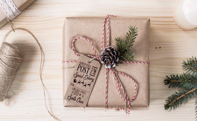 May include: A brown paper gift box tied with twine and a red and white striped ribbon. A pine sprig and a pine cone are attached to the box. A tag is attached to the box with the text "North Pole Post Special Delivery" and "Checked Twice & Sent By Santa Claus".