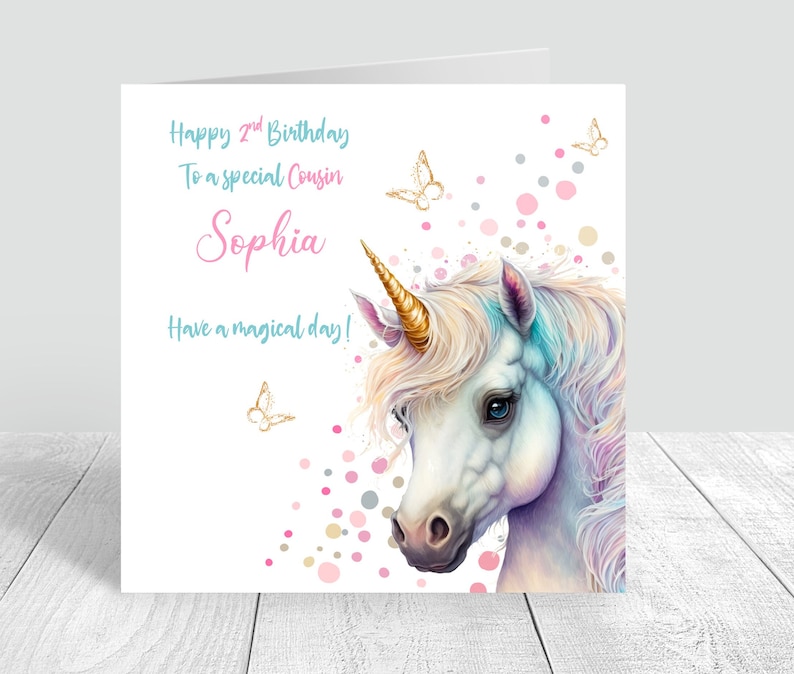 May include: A white unicorn with a gold horn and a blue and pink mane. The unicorn is surrounded by pink, gold, and gray confetti and butterflies. The text "Happy 2nd Birthday To a special Cousin Sophia Have a magical day!" is written on the card.