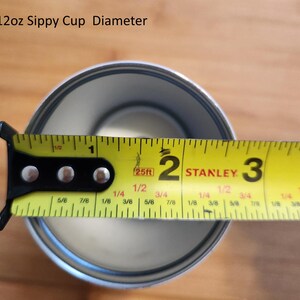 May include: A yellow measuring tape with black markings is used to measure the diameter of a silver sippy cup. The tape reads "25ft" and shows measurements in inches and fractions of an inch. The cup is 2 and 1/8 inches in diameter.
