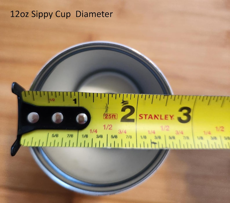 May include: A yellow measuring tape with black markings is used to measure the diameter of a silver sippy cup. The tape reads "25ft" and shows measurements in inches and fractions of an inch. The cup is 2 and 1/8 inches in diameter.
