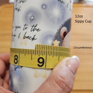 May include: A yellow measuring tape wrapped around a white ceramic sippy cup with a blue and white starry design. The tape reads 8 and 9 inches. The cup is labeled "12oz Sippy Cup" and "Circumference".