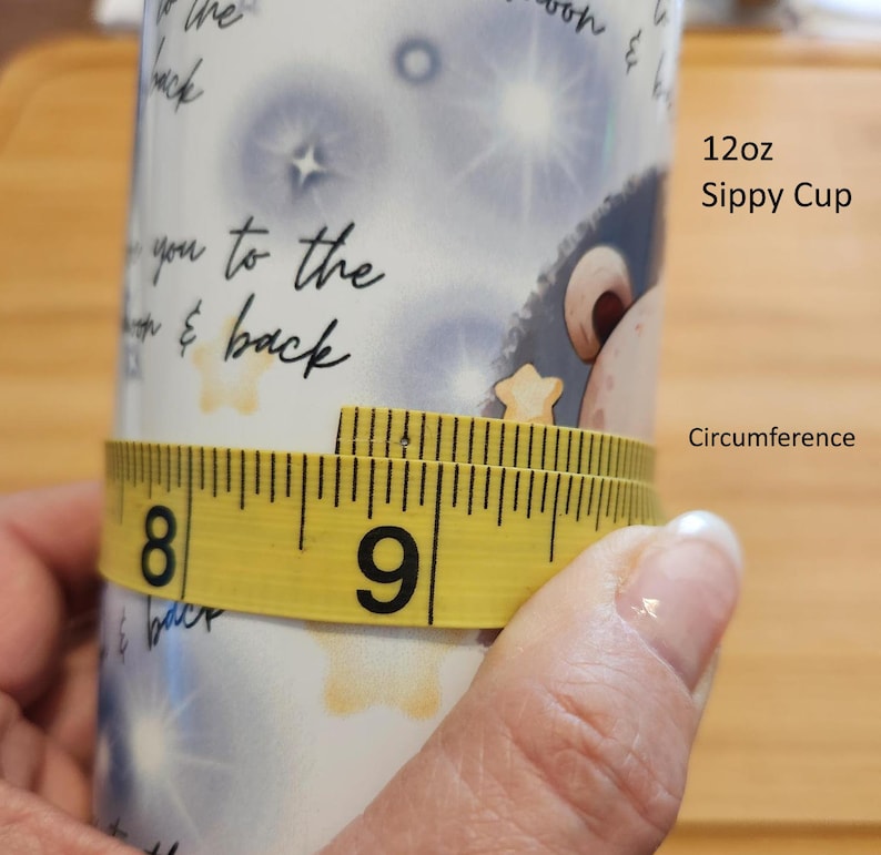 May include: A yellow measuring tape wrapped around a white ceramic sippy cup with a blue and white starry design. The tape reads 8 and 9 inches. The cup is labeled "12oz Sippy Cup" and "Circumference".