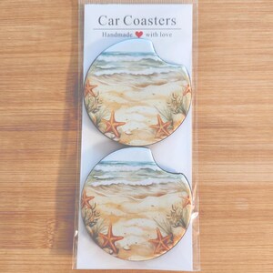 May include: Two car coasters with a beach scene design. The coasters feature a sandy beach with starfish, ocean waves, and the text "Car Coasters Handmade with love". The coasters are packaged in a clear plastic sleeve.
