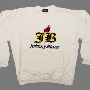 May include: Off-white long-sleeved sweatshirt featuring the text "Johnny Blaze" in black and gold. The design includes a flame and the letters "JB" in a decorative font. The sweatshirt has a round neck and ribbed cuffs.