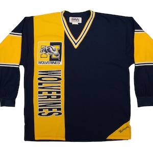 Peut inclure: Un t-shirt à manches longues avec un motif jaune et bleu. Le t-shirt a un col en V et présente un graphique d'un loup avec le mot "WOLVERINES" imprimé dessus. Le mot "WOLVERINES" est également imprimé verticalement sur le côté du t-shirt.