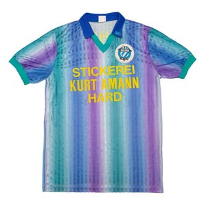 May include: A short-sleeved soccer jersey with a blue, teal, and purple striped pattern. The jersey has a blue collar and teal sleeve cuffs. The text "STICKEREI KURT AMANN HARD" is printed in yellow on the front, along with a team logo.