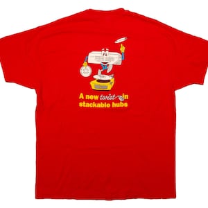 Puede incluir: Camiseta roja con un dibujo animado de un personaje de teclado que hace malabarismos con un reloj y un frisbee sobre una plataforma amarilla. El texto "A new twist in stackable hubs" está debajo.