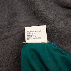 May include: A white care label with black text, including washing instructions: Machine wash cold, no bleach, hand dry, do not iron, do not dry clean. The label is attached to a teal fabric and set against a dark gray background.