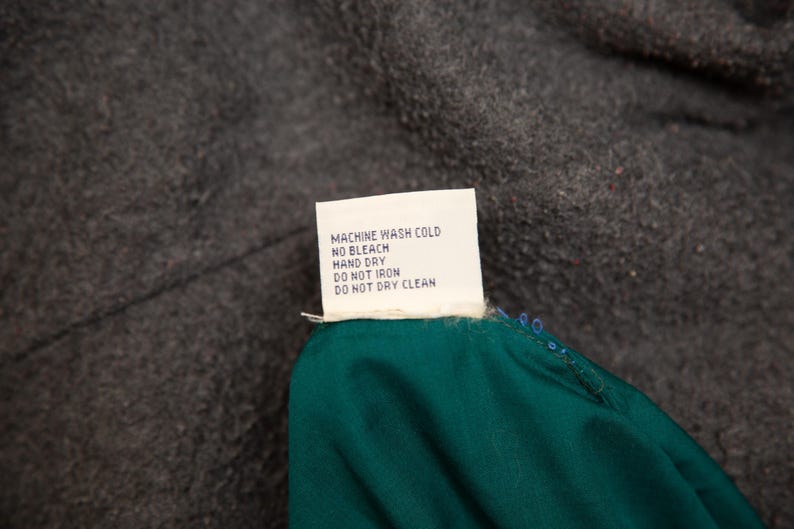 May include: A white care label with black text, including washing instructions: Machine wash cold, no bleach, hand dry, do not iron, do not dry clean. The label is attached to a teal fabric and set against a dark gray background.