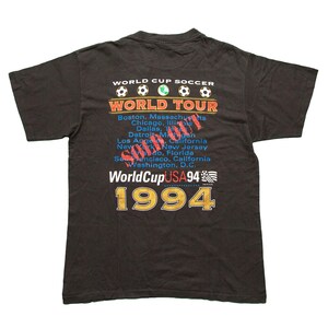 K&ouml;nnte beinhalten: Schwarzes T-Shirt mit einem Grafikdruck der World Cup Soccer World Tour 1994. Die Grafik enth&auml;lt den Text "World Cup Soccer" und "World Tour" mit einem Fu&szlig;ball-Grafik. Der Text "1994" ist in gro&szlig;en, fetten Buchstaben gedruckt.