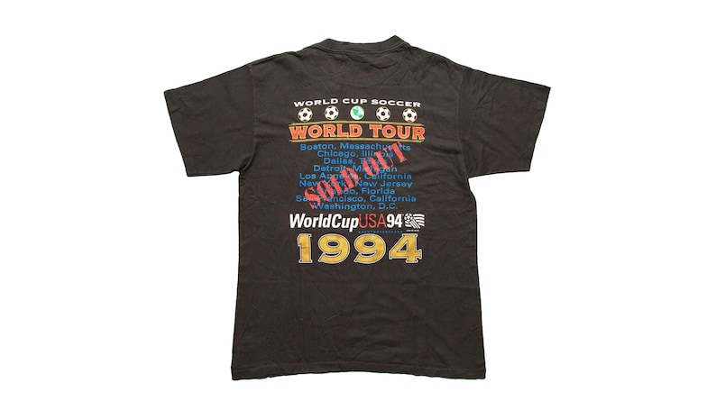 K&ouml;nnte beinhalten: Schwarzes T-Shirt mit einem Grafikdruck der World Cup Soccer World Tour 1994. Die Grafik enth&auml;lt den Text "World Cup Soccer" und "World Tour" mit einem Fu&szlig;ball-Grafik. Der Text "1994" ist in gro&szlig;en, fetten Buchstaben gedruckt.