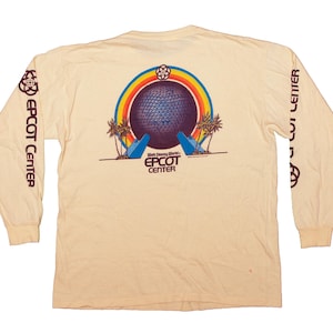 Puede incluir: Camiseta de manga larga color crema con un gráfico colorido del Centro Epcot. El diseño incluye una gran esfera, un arcoíris, palmeras y las palabras "Epcot Center". Las mangas tienen las palabras "Epcot Center" impresas verticalmente.