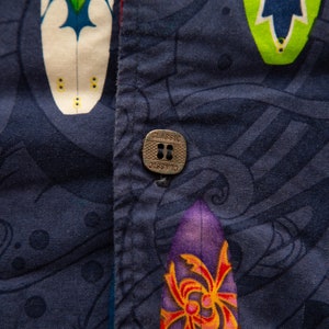 May include: Close-up of a navy blue button-down shirt with a vibrant surfboard pattern. The shirt features a metal button with the word "CLASSIC" and a red seam. The surfboards are in various colors.