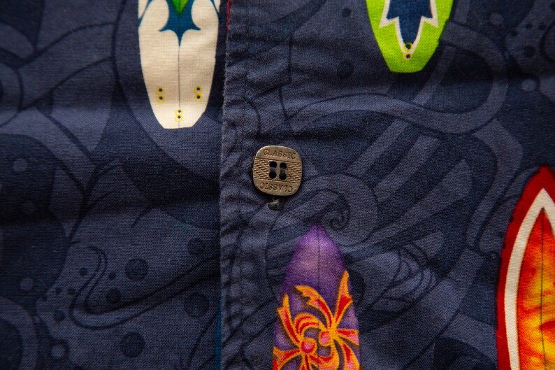 May include: Close-up of a navy blue button-down shirt with a vibrant surfboard pattern. The shirt features a metal button with the word "CLASSIC" and a red seam. The surfboards are in various colors.
