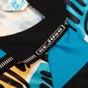 May include: Close-up of a colorful athletic jersey with a geometric pattern in black, yellow, and turquoise. The jersey has a black collar with the brand name "CE Judo" in white. A small tag is visible at the top.
