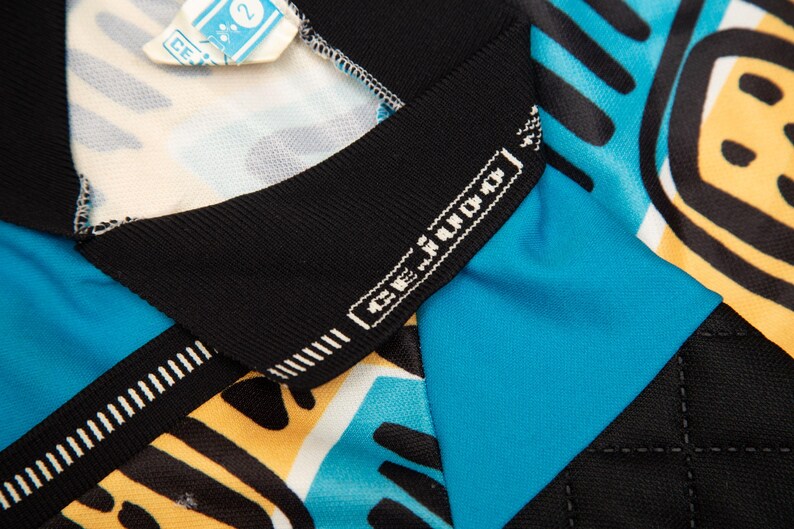 May include: Close-up of a colorful athletic jersey with a geometric pattern in black, yellow, and turquoise. The jersey has a black collar with the brand name "CE Judo" in white. A small tag is visible at the top.