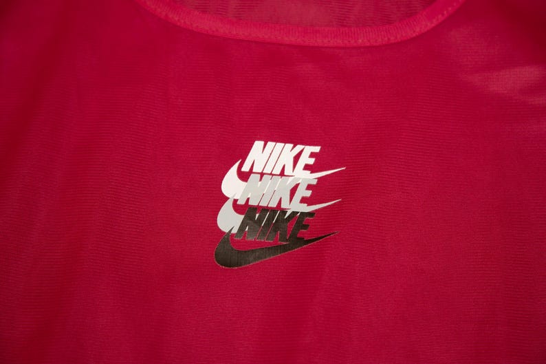 May include: A close-up of a red athletic top featuring the Nike logo. The logo is stacked three times, with the top logo in white, the middle in grey, and the bottom in black.