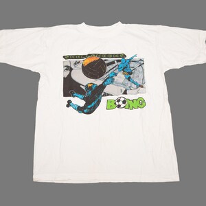 May include: A white short-sleeved t-shirt featuring a graphic print of a football scene. The design includes the text "SKULL SHOCKS" and "BOING" with a football replacing the letter O. The illustration depicts a blue player diving for a ball.