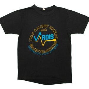 Puede incluir: Camiseta negra con un diseño circular. El diseño incluye el texto "I WAS CAUGHT ROCKIN' with VARDIS" en dorado, un rayo azul y amarillo, y "26 SEPTEMBER 1981, SHEPPERTON FILM STUDIOS" debajo.