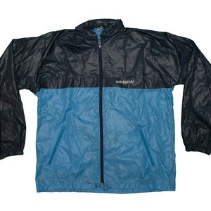 Op de afbeelding: Een vintage Adidas trainingsjack in zwart en lichtblauw. Het jack heeft een volledige ritssluiting, twee zijzakken en het Adidas-logo. Een klassiek sportkledingstuk.