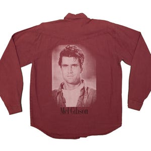 Peut inclure: Chemise à manches longues bordeaux avec une image en noir et blanc de Mel Gibson au dos. Le texte "Mel Gibson" est imprimé sous l'image. La chemise a un col boutonné et des poignets boutonnés.