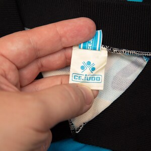 May include: Close-up of a tag with the word "CEJUDO" in a blue rectangular box. The tag is held by a hand and is attached to a black collar. The collar is part of a blue and white patterned garment.