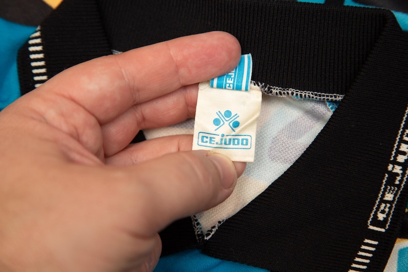 May include: Close-up of a tag with the word "CEJUDO" in a blue rectangular box. The tag is held by a hand and is attached to a black collar. The collar is part of a blue and white patterned garment.
