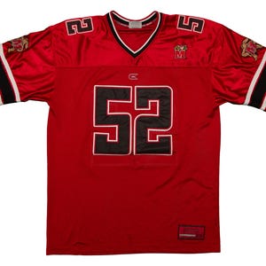 May include: A red football jersey with the number 52 in black outlined in white on the front. The jersey has a V-neck, black and white stripes on the sleeves, and a logo on the shoulders. The jersey is made of a shiny material.