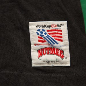 K&ouml;nnte beinhalten: Schwarzes T-Shirt mit einem wei&szlig;en Patch mit dem Text "World Cup USA 94" und einer Fu&szlig;ballgrafik. Der Patch enth&auml;lt auch den Text "Nutmeg" und "Made in U.S.A."