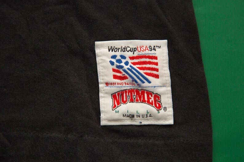 K&ouml;nnte beinhalten: Schwarzes T-Shirt mit einem wei&szlig;en Patch mit dem Text "World Cup USA 94" und einer Fu&szlig;ballgrafik. Der Patch enth&auml;lt auch den Text "Nutmeg" und "Made in U.S.A."
