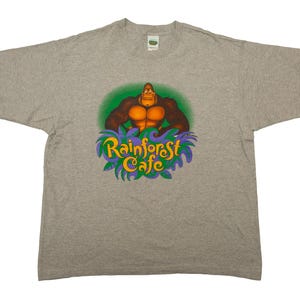 May include: A light gray t-shirt featuring a cartoon gorilla with orange chest muscles, set against a green circle. The text "Rainforest Cafe" is in yellow and purple, surrounded by green leaves.