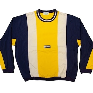 May include: A long-sleeved IZOD sweatshirt in a color block design. The shirt features a yellow center panel, flanked by a cream-colored panel, and navy blue sleeves. The collar is trimmed with white and navy blue stripes. The IZOD logo is centered.