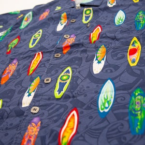 May include: A navy blue button-down shirt with a vibrant pattern of colorful surfboards. The surfboards are in various colors, including red, yellow, green, and orange, and are set against a background of swirling blue designs. The shirt has a classic collar and buttoned cuffs.