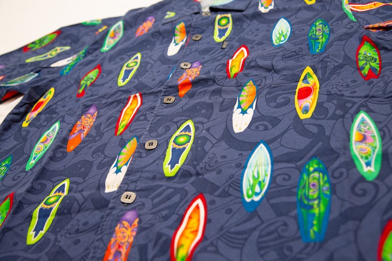 May include: A navy blue button-down shirt with a vibrant pattern of colorful surfboards. The surfboards are in various colors, including red, yellow, green, and orange, and are set against a background of swirling blue designs. The shirt has a classic collar and buttoned cuffs.