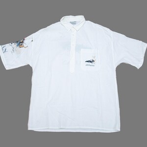 Könnte beinhalten: Ein weißes kurzärmeliges Poloshirt mit Knopfleiste, Kragen und Brusttasche. Das Hemd zeigt einen kleinen grafischen Aufdruck auf der Tasche und eine größere, detaillierte grafische Illustration in Blau- und Weißtönen auf dem rechten Ärmel.