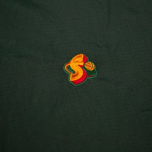 May include: Dark green fabric with a small, embroidered logo. The logo features a stylized "S" in yellow and orange, with a basketball design and green and red accents. The fabric has a subtle textured pattern.