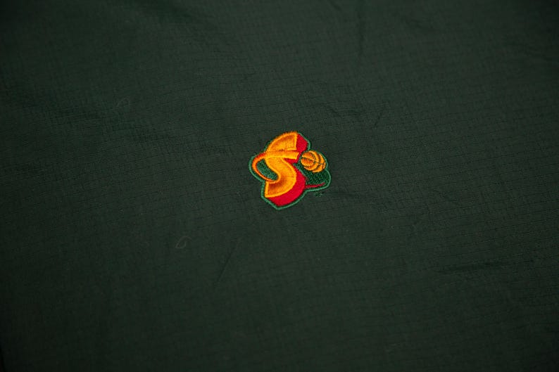 May include: Dark green fabric with a small, embroidered logo. The logo features a stylized "S" in yellow and orange, with a basketball design and green and red accents. The fabric has a subtle textured pattern.