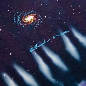 May include: Close-up of a fabric with a cosmic design. A spiral galaxy in shades of orange, yellow, and blue is surrounded by white stars on a dark blue background. The text "&copy;1992 Liquid Blue" and a signature are visible.