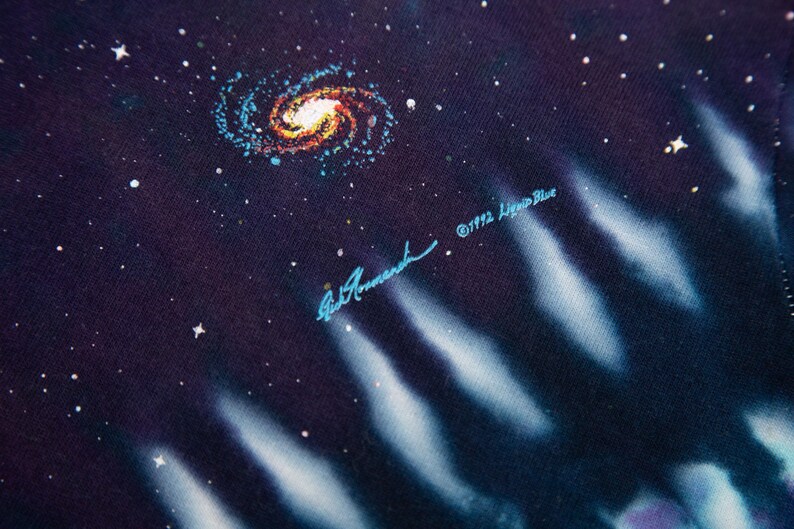 May include: Close-up of a fabric with a cosmic design. A spiral galaxy in shades of orange, yellow, and blue is surrounded by white stars on a dark blue background. The text "&copy;1992 Liquid Blue" and a signature are visible.