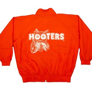 May include: Bright orange jacket with a white logo that says "HOOTERS" and a graphic of an owl. The jacket has a zipper, elastic cuffs, and a drawstring hem. The jacket is made of a lightweight material.