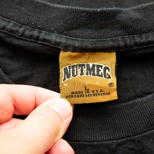 K&ouml;nnte beinhalten: Ein schwarzes T-Shirt mit einem goldenen Etikett, auf dem "Nutmeg Mills" und "Made in U.S.A. For care see reverse." steht.