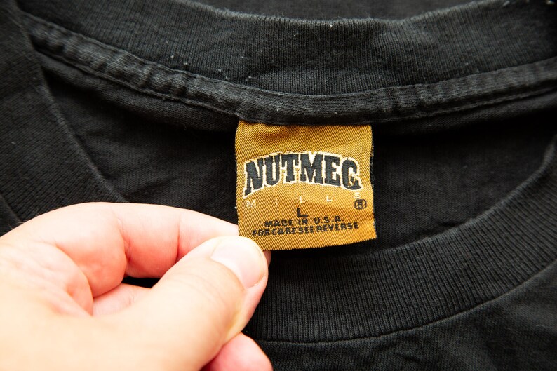K&ouml;nnte beinhalten: Ein schwarzes T-Shirt mit einem goldenen Etikett, auf dem "Nutmeg Mills" und "Made in U.S.A. For care see reverse." steht.