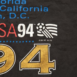 K&ouml;nnte beinhalten: Schwarzes T-Shirt mit dem Text "USA 94" in Rot und Wei&szlig;, einer Fu&szlig;ballgrafik und dem Text "&copy;1991 WC 94/ISL". Die Zahlen "94" sind in Gold.