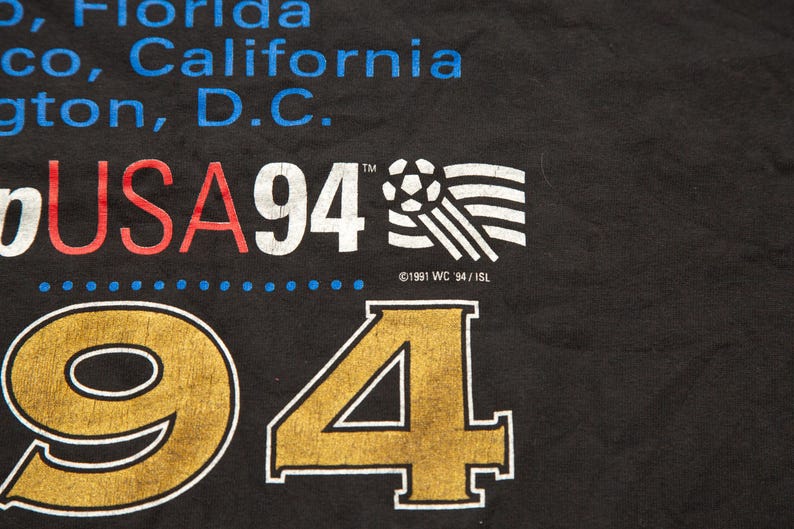 K&ouml;nnte beinhalten: Schwarzes T-Shirt mit dem Text "USA 94" in Rot und Wei&szlig;, einer Fu&szlig;ballgrafik und dem Text "&copy;1991 WC 94/ISL". Die Zahlen "94" sind in Gold.