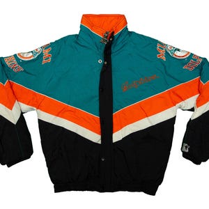May include: A teal, orange, white, and black jacket with the Miami Dolphins logo on the sleeves. The jacket has a high collar and a button-up front. The word "Dolphins" is embroidered on the front.