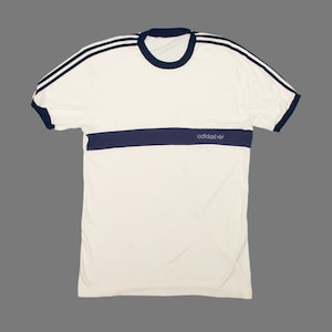 May include: Cream-colored t-shirt with navy blue accents. Features navy blue stripes on the shoulders and a horizontal stripe across the chest. The Adidas logo is visible on the chest stripe. The shirt has a crew neck and short sleeves.