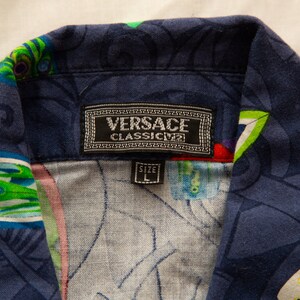 May include: Close-up of a navy blue button-down shirt with a colorful, abstract pattern. The shirt collar features a label with the "VERSACE CLASSIC V2" logo and a size "L" tag.
